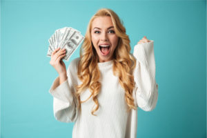 Portrait of an excited young blonde woman in sweater showing money banknotes isolated over blue background