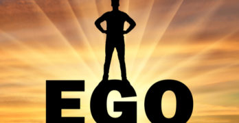Silhouette of a man with a crown on his head standing on the word ego against the backdrop of a sunset. Concept of selfishness