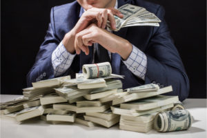 Man seated in a business suit holding Money in his hands with a pile of money on the table in front of him.